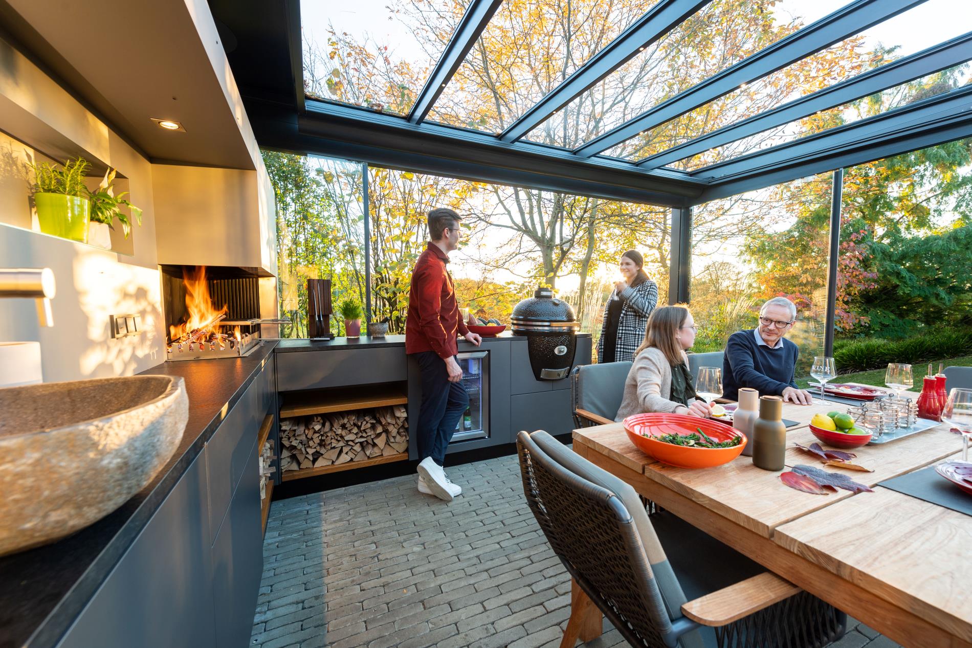 Antracitszürke kültéri konyha fatüzelésű beépített grillel, kamado grillel és hűtővel, modern Solarlux üvegterasz alatt, terített étkezőasztallal és beszélgető társasággal.