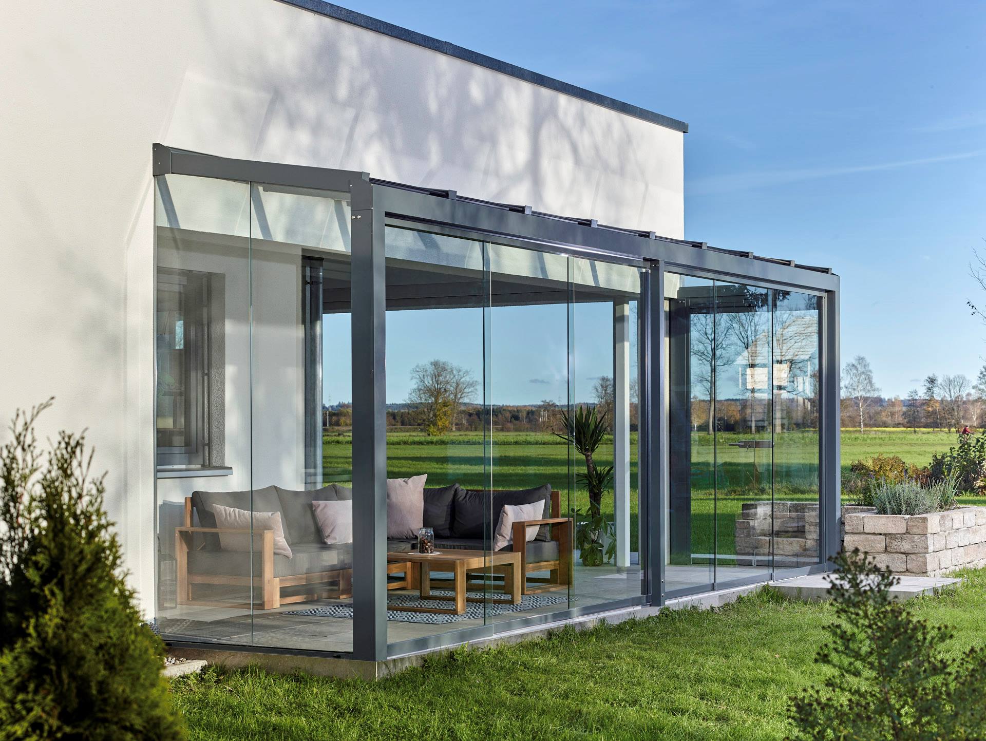 Antracitszürke Solarlux Alerio üvegterasz eltolható üvegfalakkal, alatta modern kerti bútorokkal és kanapéval, háttérben zöld mezővel.