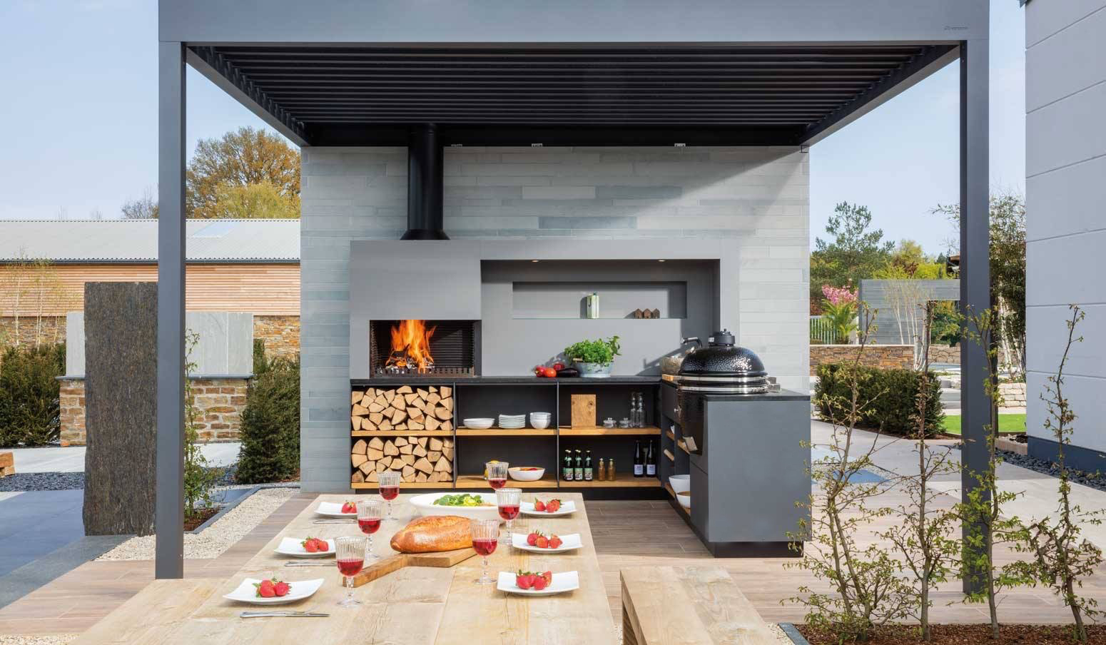 L-alakú Freiluft kültéri konyha fatüzelésű beépített grillel, tűzifatárolóval és kamado grillel, egy modern lamellás pergola alatt, terített kerti étkezőasztallal.